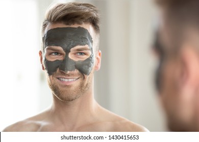 Handsome man looks at himself through the mirror with a cosmetic mask on his face
