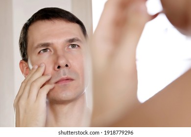 Handsome Man Looks At His Reflection In The Mirror Touching His Face. Male Beauty, Health And Skin Care Concept