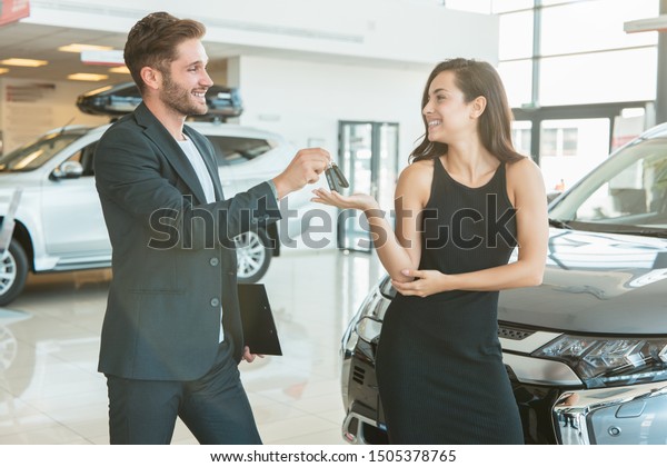 handsome man manager giving car keys to beautiful\
woman client standing near brand new car after succesful deal in\
dealership center looking\
happy