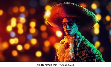 Handsome man in Mexican sombrero and traditional costume with skull makeup on his face. Festive background with colorful lights and Copy Space. Dia de los muertos. Day of The Dead. Halloween. - Shutterstock ID 2205375503