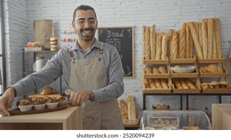 Handsome man with moustache standing in bakery shop offering muffins and smiling at camera with bread displayed in the background on wooden shelves - Powered by Shutterstock