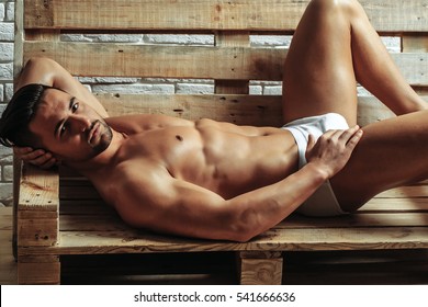 Handsome man or muscular macho, bodybuilder, with sexy, muscle torso, body, with six packs and abs in sexi underpants on wooden pallet sofa on white brick wall background - Shutterstock ID 541666636