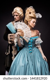 Handsome Man Near Victorian Woman In Wig Holding Fan On Black 