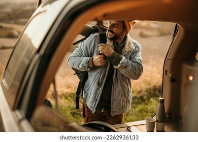 A handsome man packed his backpack, put it on a shoulder, and took it out from a car truck, getting ready for a nature walk and hike. - Powered by Shutterstock