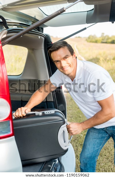 handsome man packing suitcase into luggage boot\
of car in field and looking at\
camera