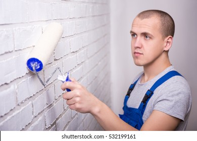 Handsome Man Painter In Work Wear Painting White Brick Wall With Paint Roller