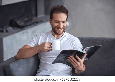 Handsome man is reading a magazine, holding a cup and smiling while resting at home - Shutterstock ID 2370102277
