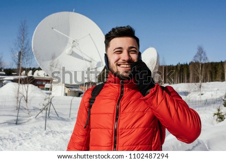 Handsome man in red jacket talking on the cell phone while standing near the radio observatory with big satellite dishes in the forest in winter. Norway