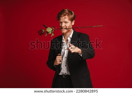 Handsome man with red lipstick marks on his face pointing finger at camera while holding rose in his mouth isolated over red studio wall