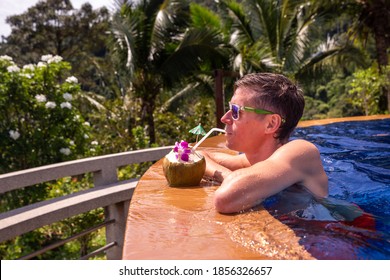 Handsome man relaxes by the outdoor pool and drinks a coconut with palm trees in the background. Summer concept.