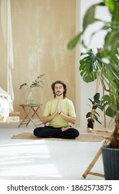Handsome Man Sitting In Lotus Pose Holding Arms In Namaste, Meditation Teacher Practicing Concentration At Home With Green Plants