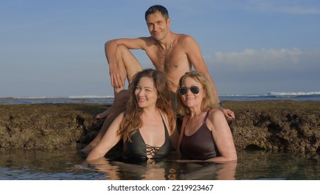 Handsome Man Sitting On A Reef Stone And Two Women Bathing In Ocean Water Posing And Smiling For The Picture On Beach At Sunset. Happy Time For A Family Spending Time Together. Soft Focus.