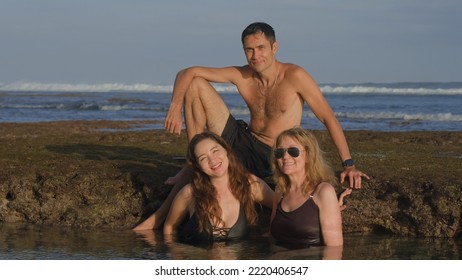 Handsome Man Sitting On A Reef Stone And Two Smiling Women Bathing In Ocean Water Posing And Smiling For The Picture On Beach At Sunset. Happy Time For A Family Spending Time Together. Soft Focus.