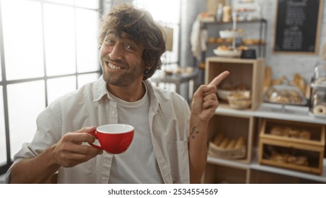 Handsome man smiling and holding a red coffee cup while pointing inside a cozy bakery with fresh pastries on display shelves - Powered by Shutterstock