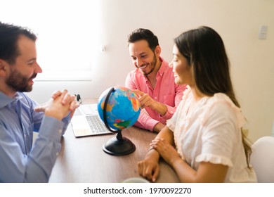 Handsome Man Smiling And Pointing A Foreign Country On A Globe At The Travel Agency. Couple Choosing Their Next Destination Abroad