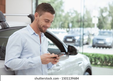 Handsome Man Smiling, Texting On His Mobile Phone Standing Near New Automobile At Dealership Salon. Cheerful Man Buying New Auto, Using Smart Phone