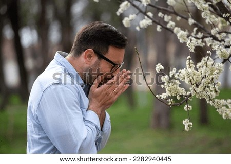 Handsome man sneezing beside blooming tree in spring outdoors. Allergy pollen symptoms concept