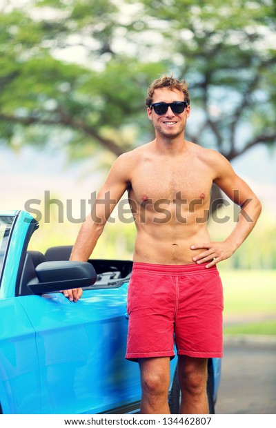 Handsome man with sports car. Well built fit\
young male model standing shirtless leaning on the door of a blue\
sports car smiling at the\
camera.