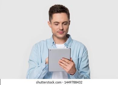 Handsome man standing scrolling and typing on electronic tablet device. Happy male holding gadget using popular application, typing message on screen. Photoshoot at studio isolated on gray background