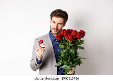 Handsome man in suit, showing engagement ring and looking romantic at camera, standing with red roses over white background. Valentines day and love concept