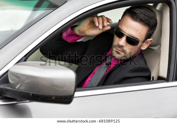 Handsome man in\
suit and sunglasses, sitting in silver/grey car looking through\
open car window in side\
mirror.