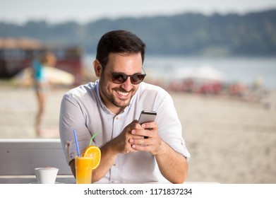 Handsome Man With Sunglasses Drinking Coffee And Juice And Looking At Mobile Phone In Cafe On Beach. Enjoying Morning Beside River