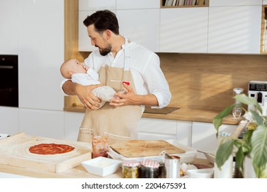 Handsome Man Talking With Lovely Baby Holding In Arms. Adorable Father In Aprons Cooking Preparing Homemade Pizza Dressing Dough Applying Tomato Sauce.