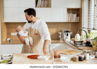 Handsome Man Talking With Lovely Baby Holding In Arms. Adorable Father In Aprons Cooking Preparing Homemade Pizza Dressing Dough Applying Tomato Sauce.