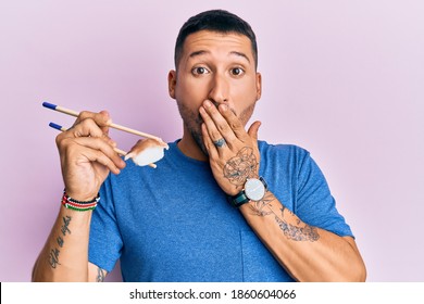 Handsome man with tattoos eating eel sushi using chopsticks covering mouth with hand, shocked and afraid for mistake. surprised expression  - Powered by Shutterstock