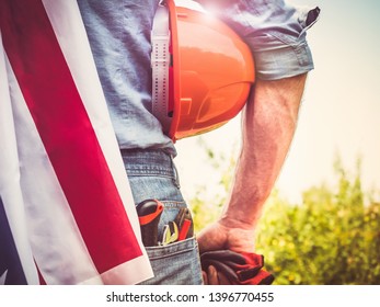 Handsome man with tools, holding an American Flag, standing against a background of green trees and the rays of the setting sun. View from the back, close-up. Concept of work and employment - Shutterstock ID 1396770455