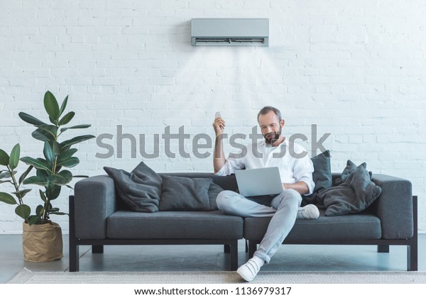 handsome man turning on air\
conditioner with remote control while using laptop on sofa at\
home