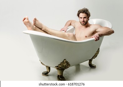 Handsome man or unshaven caucasian macho with stylish haircut, hair, with naked, sexy muscular torso and arms with biceps, triceps sitting in classic bath, tub barefoot on white background