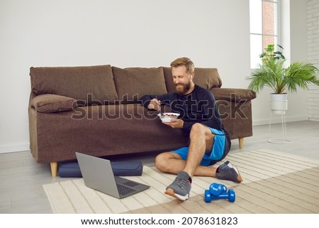 Handsome man using laptop during workout break. Sporty guy regaining energy after active indoor weightloss physical exercise, sitting by sofa at home, holding bowl, eating healthy food, watching video