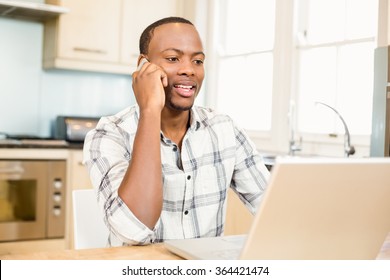 Handsome Man Using Laptop And Having A Phone Call In The Kitchen. Working From Home In Quarantine Lockdown. Social Distancing Self Isolation