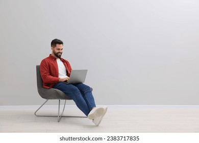 Handsome man using laptop while sitting in armchair near light grey wall indoors, space for text - Powered by Shutterstock