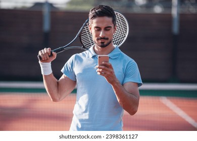 Handsome man is using a smart phone while playing tennis on tennis court outdoors - Powered by Shutterstock