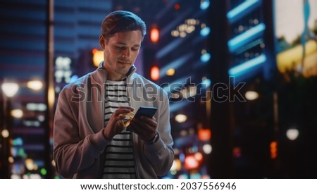 Handsome Man Using Smartphone Walking Through Night City Full of Neon Colors and Entertainment. Stylish Young Man Using Mobile Phone, Posting on Social Media, Online Shopping, Texting on Dating App