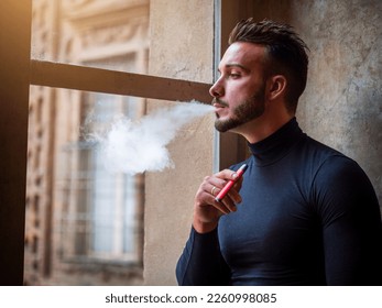 Handsome man vaping, smoking e-cigarette by a window - Powered by Shutterstock