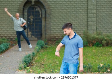 Handsome Man Is Waiting For Friend On Street And Looks At His Watch, Behind Him A Friend Waves His Hand To Him Leaving The House