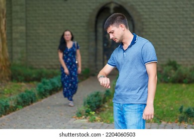 Handsome Man Is Waiting For Girl On Street And Looks At His Watch, Behind Him A Young Woman In A Dress Goes To Him Leaving The House