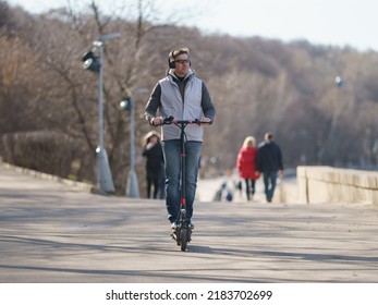 Handsome Man Walking In Moscow Public Park In Sunny Spring Day. He Is Well-dressed, Put On Sunglasses. His Clothes Is In Gray Colors. He Riding Motor Scooter And Admiring View. Healthy Lifestyle