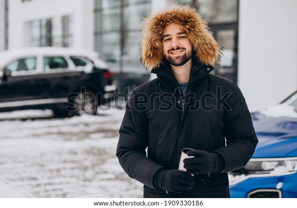Handsome man in warm jacket standing by car\
covered with snow and drinking\
coffee