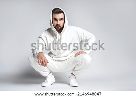 Handsome man wearing blank white hoodie and pants sitting on light gray background