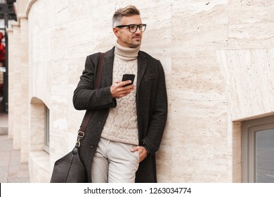 Handsome man wearing a coat walking outdoors, holding mobile phone