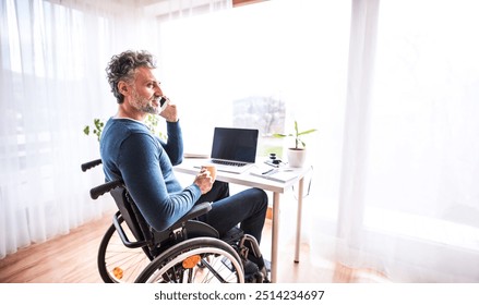 Handsome man in wheelchair working from home office, making phone call. Telecommuting and home office.