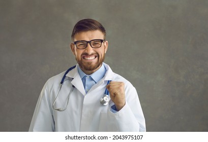 Handsome Man In White Medical Lab Coat Looking At Camera, Smiling And Fist Pumping Celebrating Professional Success. Happy Young Male Nurse Or Doctor Excited About Curing Patient Or Getting Promotion