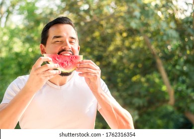 Handsome Man In White Shirt Biting Juicy Slice Of Red Watermelon Outdoors. Cheerful Adult Man Eating Watermelon On Picnic. Green Leaves In Background. Healthy Food, Freshness And Enjoying Life Concept