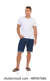 Handsome Man In White Shirt, Jeans Shorts And Black Sandals Standing With Hand In Pocket And Looking Away. Full Length Studio Shot Isolated On White.