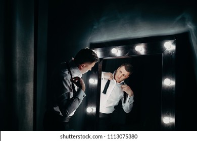 A Handsome Man In A White Shirt Looks In The Dressing Mirror And Puts On A Bow Tie In Dark Room