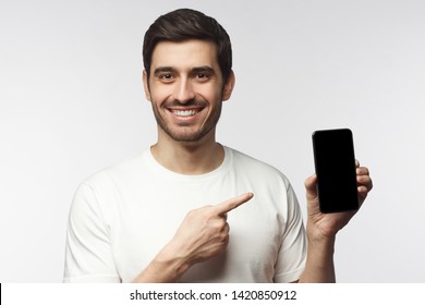 Handsome Man In White T-shirt Presenting Smart Phone And Pointing With Finger At Blank Black Screen With Copy Space, Isolated On Gray Background